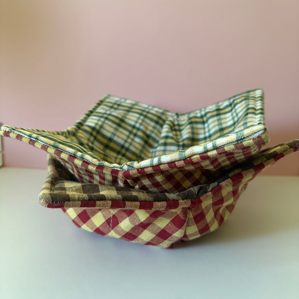Two rustic colored plaid or checkered fabric bowls stacked on a white surface with a pink background