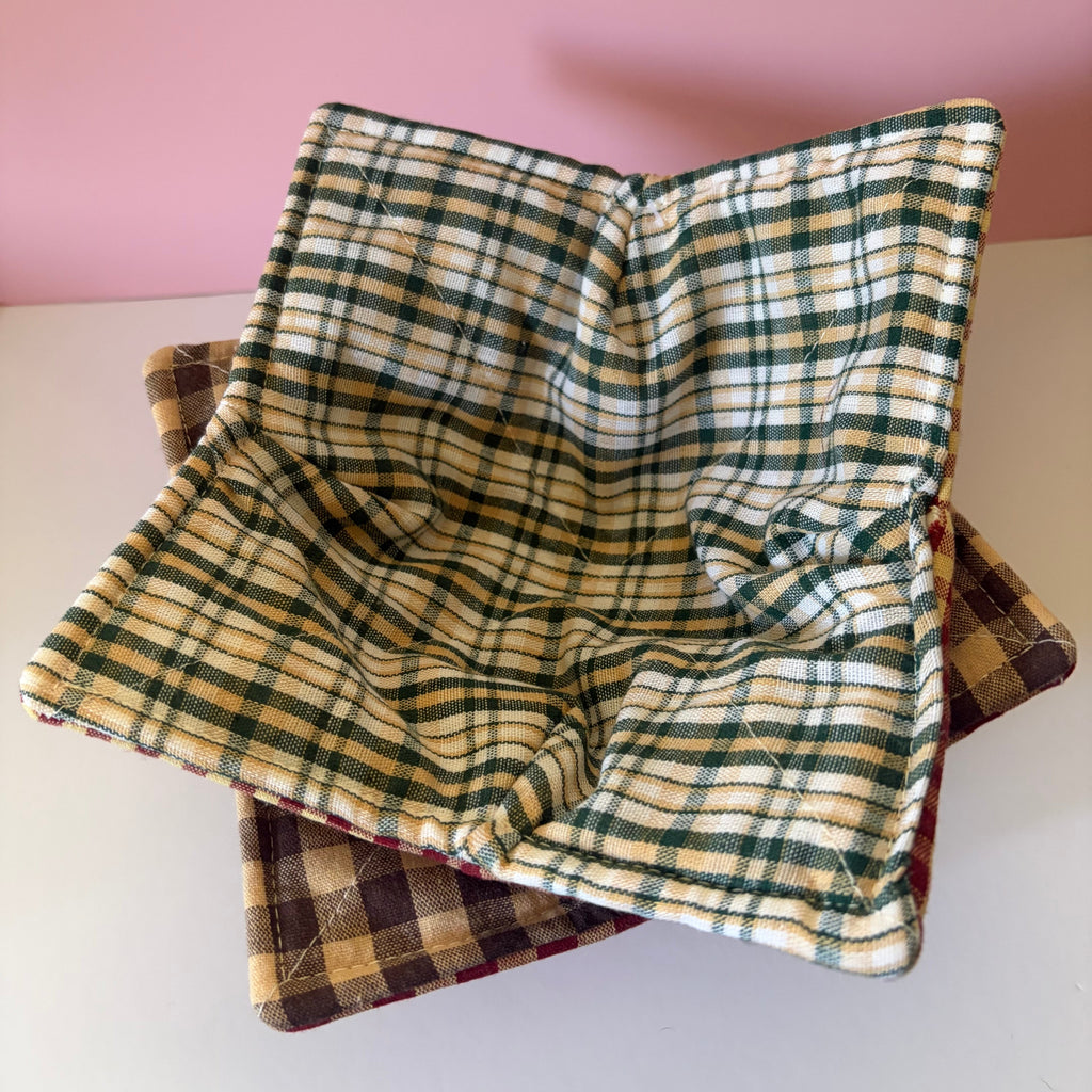 Two rustic colored plaid or checkered fabric bowls stacked on a white surface with a pink background