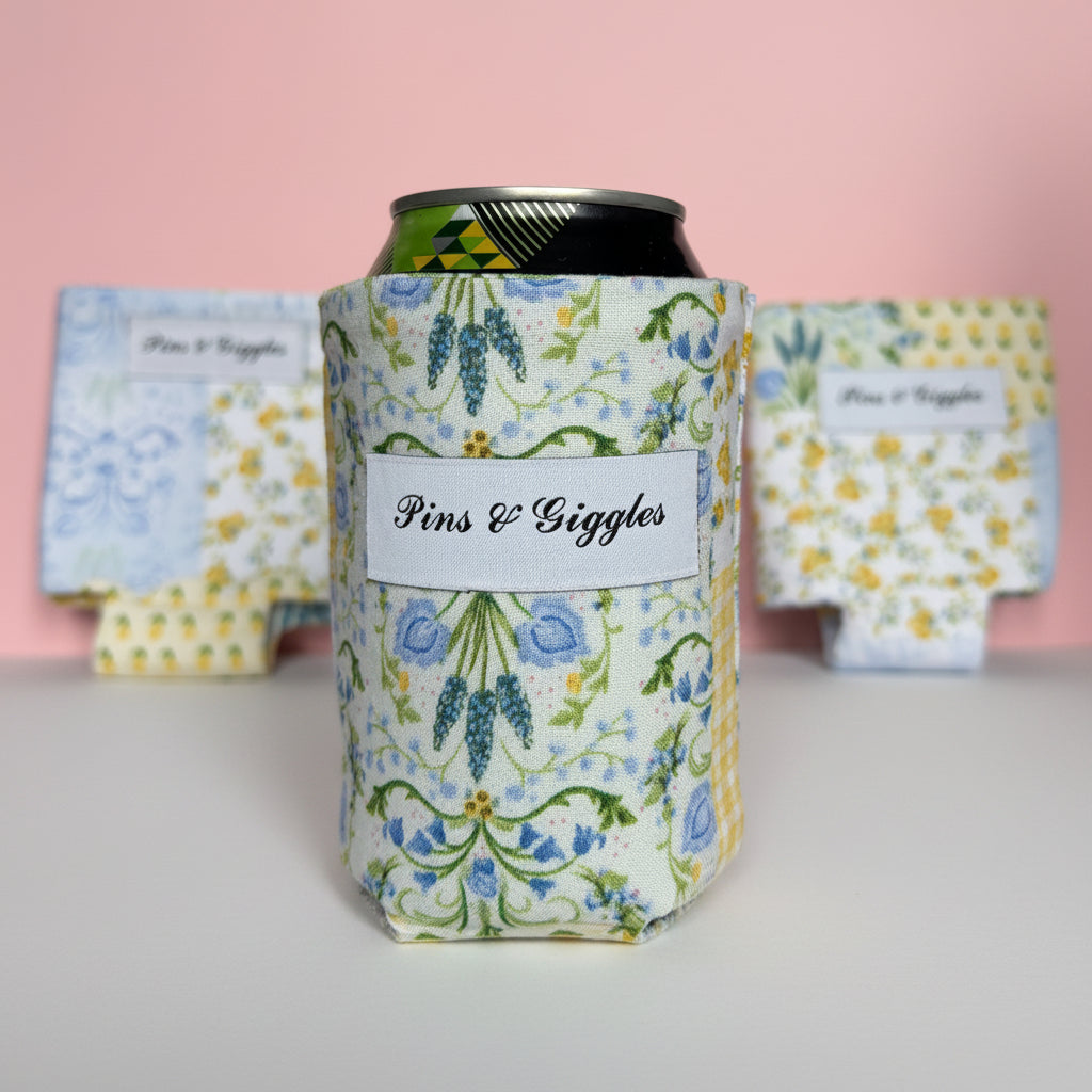 Floral can cooler with 'Pins & Giggles' branding on a pink background with other can coozies behind the main one, displaying the varying patterns.